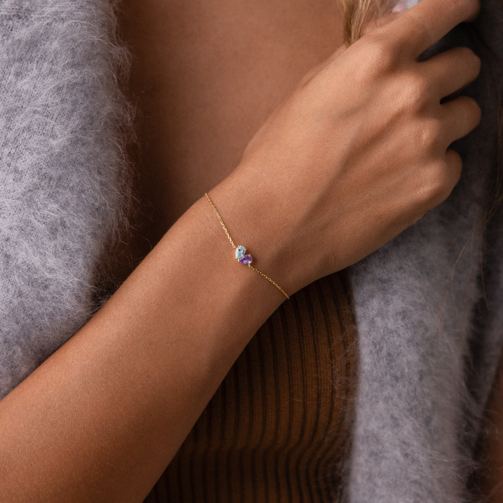 The Toi et Moi Teardrop Heart Bracelet, featuring a gold teardrop heart and purple gemstone, adorns a woman's wrist, styled with a brown top and grey sweater.
