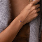 The Toi et Moi Teardrop Heart Bracelet, featuring a gold teardrop heart and purple gemstone, adorns a woman's wrist, styled with a brown top and grey sweater.