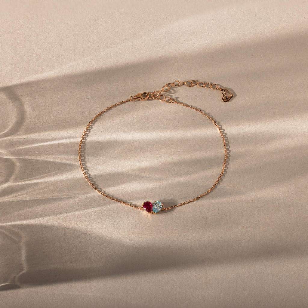 The Toi et Moi Teardrop Heart Bracelet in Rose Gold features a red and a clear gemstone, elegantly displayed on a beige surface with soft shadows.