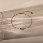The Toi et Moi Teardrop Heart Bracelet in Rose Gold features a red and a clear gemstone, elegantly displayed on a beige surface with soft shadows.
