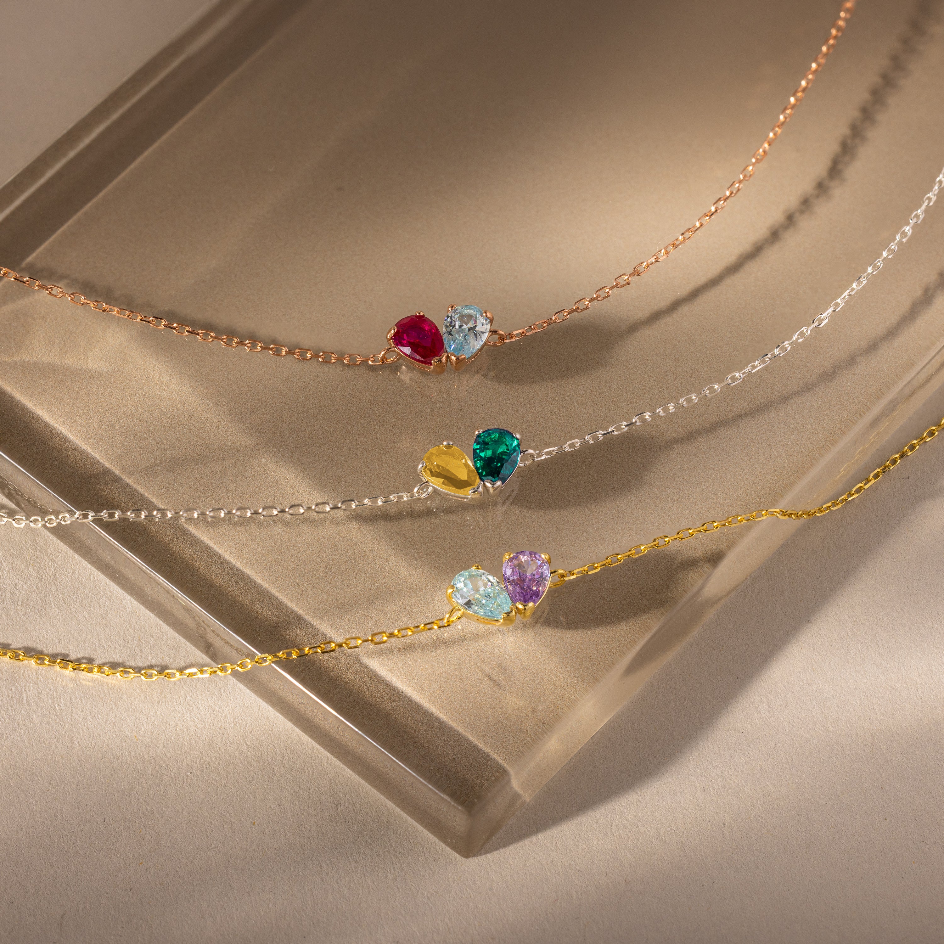 The Toi et Moi Teardrop Heart Bracelet features delicate chains and colorful teardrop heart pendants, elegantly displayed on a beige glass surface.