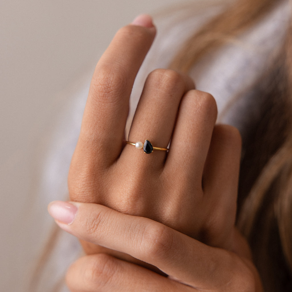 A hand wears the Teardrop Birthstone & Pearl Ring, featuring a black stone and a small pearl on the ring finger for an elegant jewelry accent.