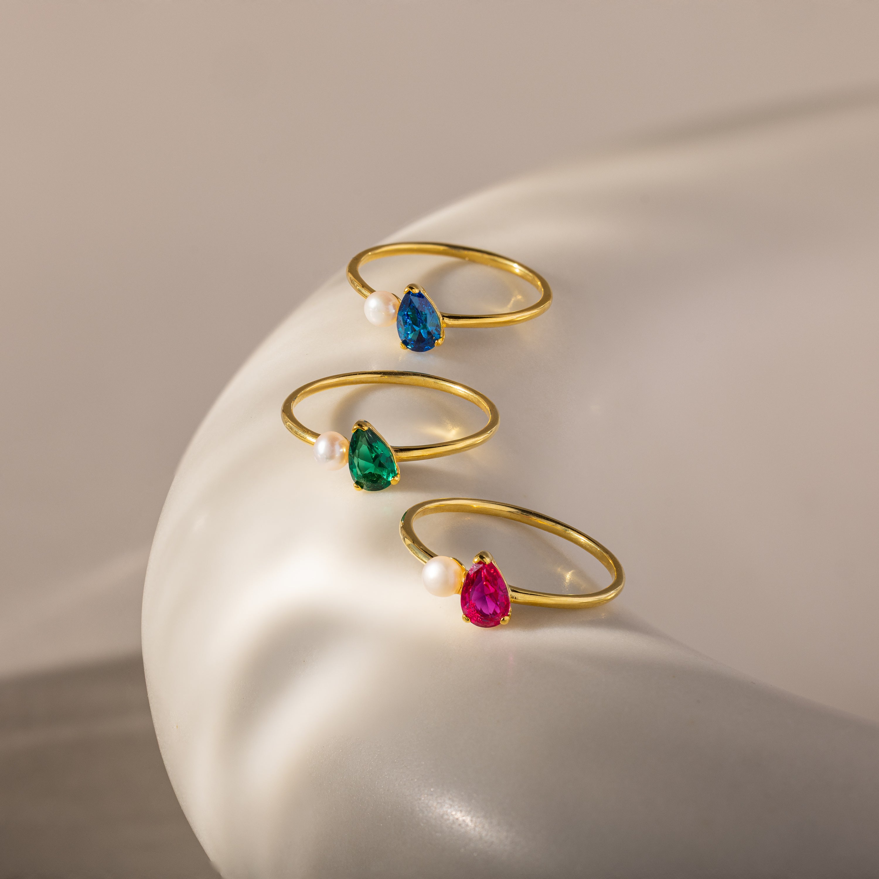 Three gold rings featuring colorful teardrop gemstones and pearls, including the striking Teardrop Birthstone & Pearl Ring, are displayed on a smooth white surface—ideal for lovers of elegant jewelry.