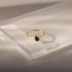 The Teardrop Onyx & Pearl Ring is an elegant gold piece featuring a black onyx teardrop birthstone and a white pearl set on a clear, angled surface.