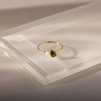 The Teardrop Peridot & Pearl Ring, featuring a teardrop peridot birthstone and a delicate white pearl, is displayed on clear acrylic—an elegant and exquisite jewelry piece.