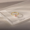 The Teardrop Pink Tourmaline & Pearl Ring, featuring a pink tourmaline stone and a delicate white pearl, rests atop a clear rectangular surface—an elegant jewelry piece with lasting charm.