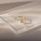 The Teardrop Pink Tourmaline & Pearl Ring, featuring a pink tourmaline stone and a delicate white pearl, rests atop a clear rectangular surface—an elegant jewelry piece with lasting charm.