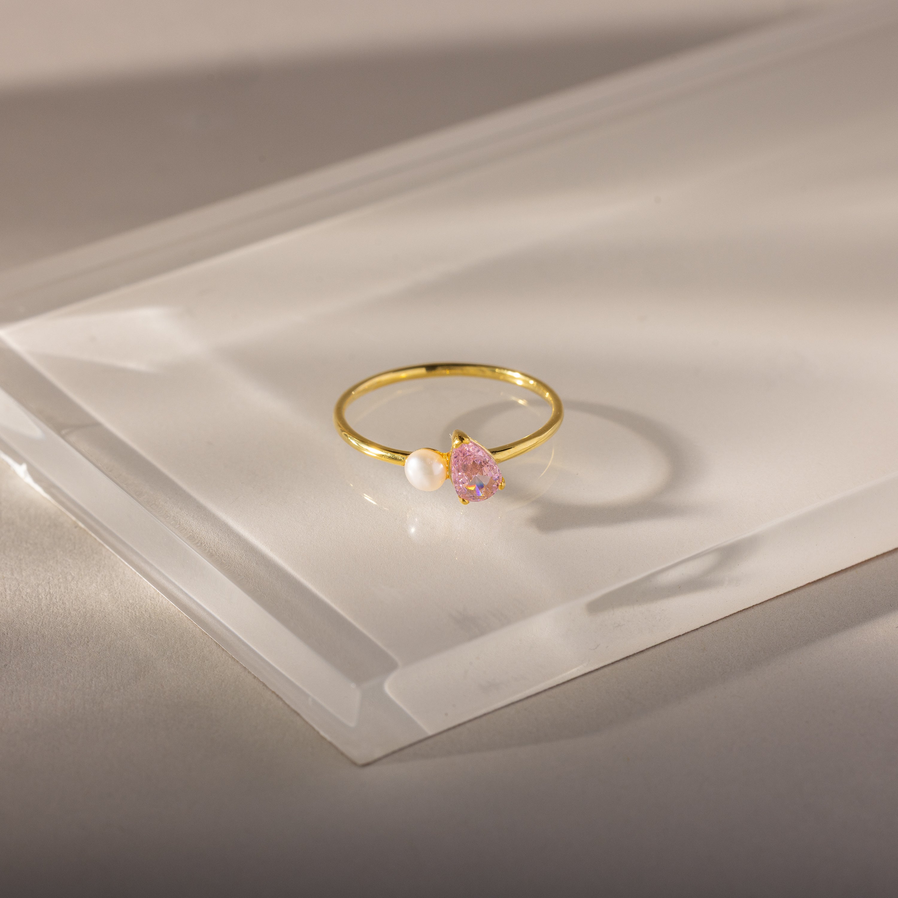 The Teardrop Pink Tourmaline & Pearl Ring, featuring a pink tourmaline stone and a delicate white pearl, rests atop a clear rectangular surface—an elegant jewelry piece with lasting charm.