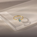 The Teardrop Aquamarine & Pearl Ring features a blue aquamarine gemstone and a pearl in an elegant design, showcased on a clear, angled surface.