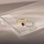 The Teardrop Amethyst & Pearl Ring features a gold band with a purple amethyst teardrop and a small white pearl, displayed on clear acrylic.