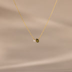 The Teardrop Peridot & Pearl Necklace, featuring a small green peridot and a white pearl pendant, rests on a soft beige background—a timeless gift for any occasion.