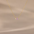 The Teardrop Alexandrite & Pearl Necklace features a gold chain with a small pearl and a purple teardrop gemstone pendant. A timeless gift for special occasions, shown here on a beige background.