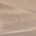 The Teardrop Alexandrite & Pearl Necklace features a gold chain with a small pearl and a purple teardrop gemstone pendant. A timeless gift for special occasions, shown here on a beige background.