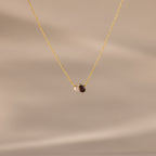 The Teardrop Garnet & Pearl Necklace features a small white pearl and a deep red garnet pendant on a delicate gold chain, making it an elegant milestone gift. Displayed on a beige background.