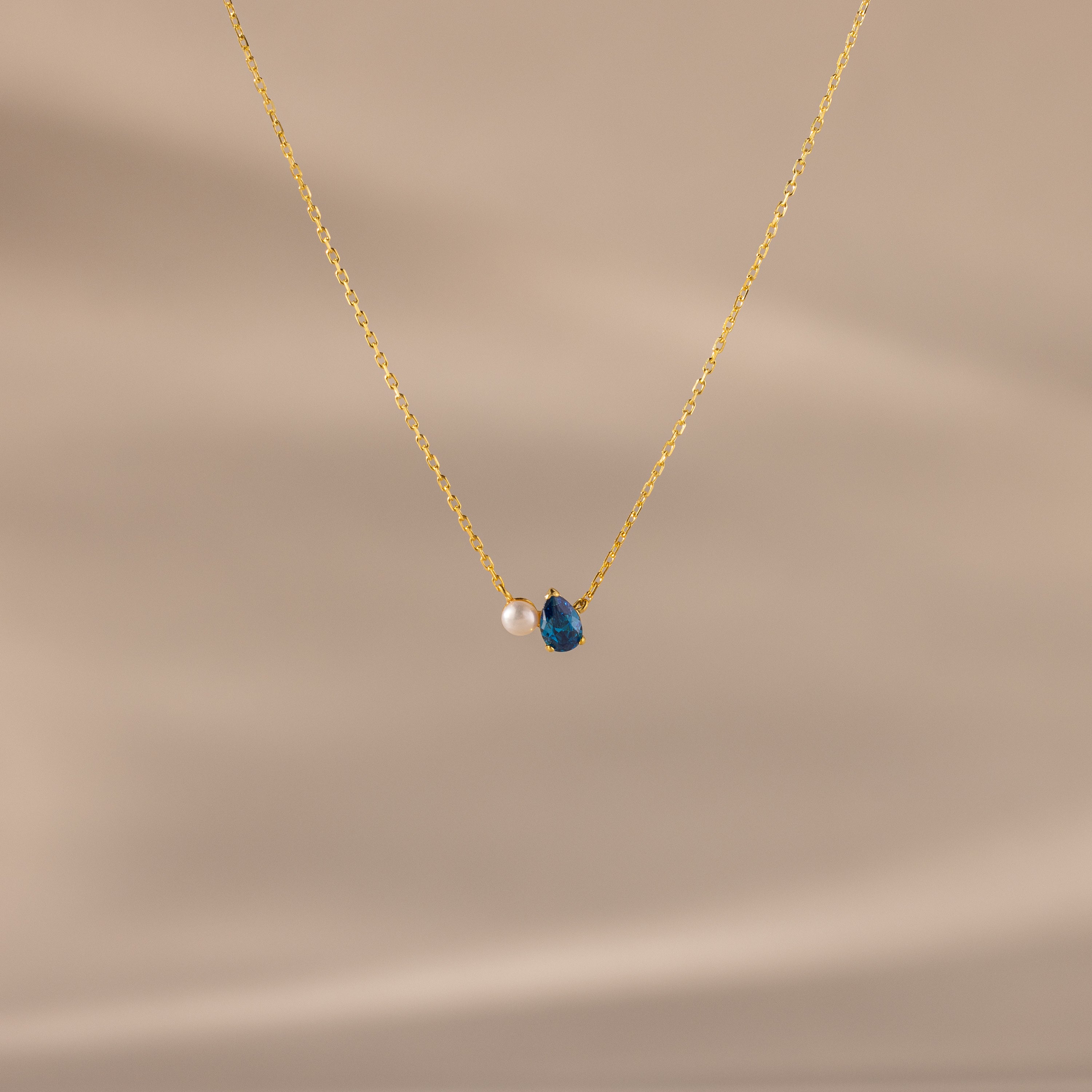 Teardrop Blue Zircon & Pearl Necklace with a small pearl and blue zircon pendant, ideal as a milestone gift, showcased on a soft beige background.