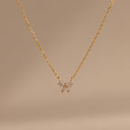 The Diamond Bow Necklace features a delicate gold-plated chain with a symmetrical bow pendant set with sparkling cubic zirconia, displayed on a soft beige background.