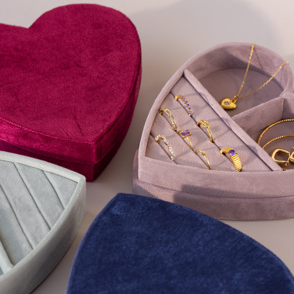 Assorted velvet heart-shaped jewelry boxes in red, blue, and mint green, with lavender box open to display gold rings, bracelet, and heart pendant necklace.