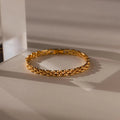 A Strap Bracelet with a textured link design rests on a beige surface in soft, shadowed lighting, highlighting it as a bold statement piece.