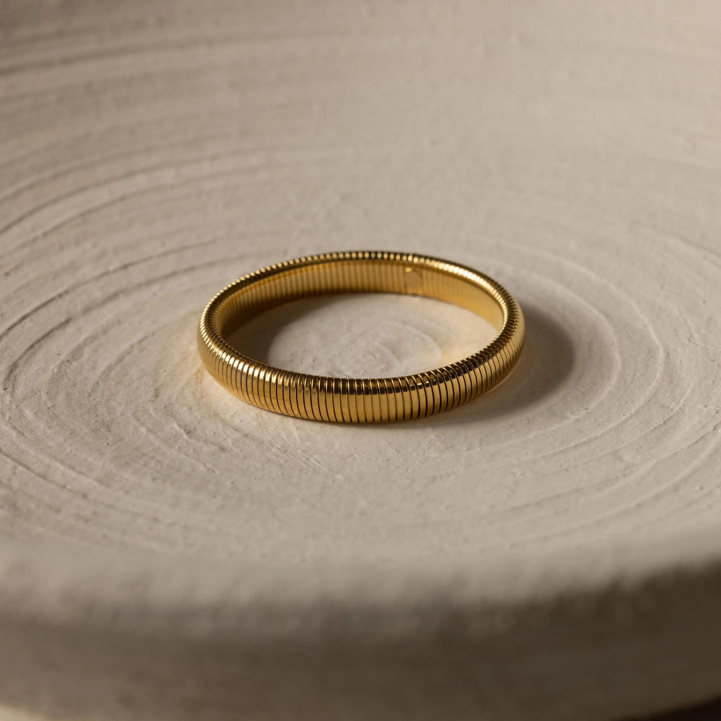 The Thick Snake Chain Bangle, featuring a bold ribbed design, is displayed on a light, textured surface.