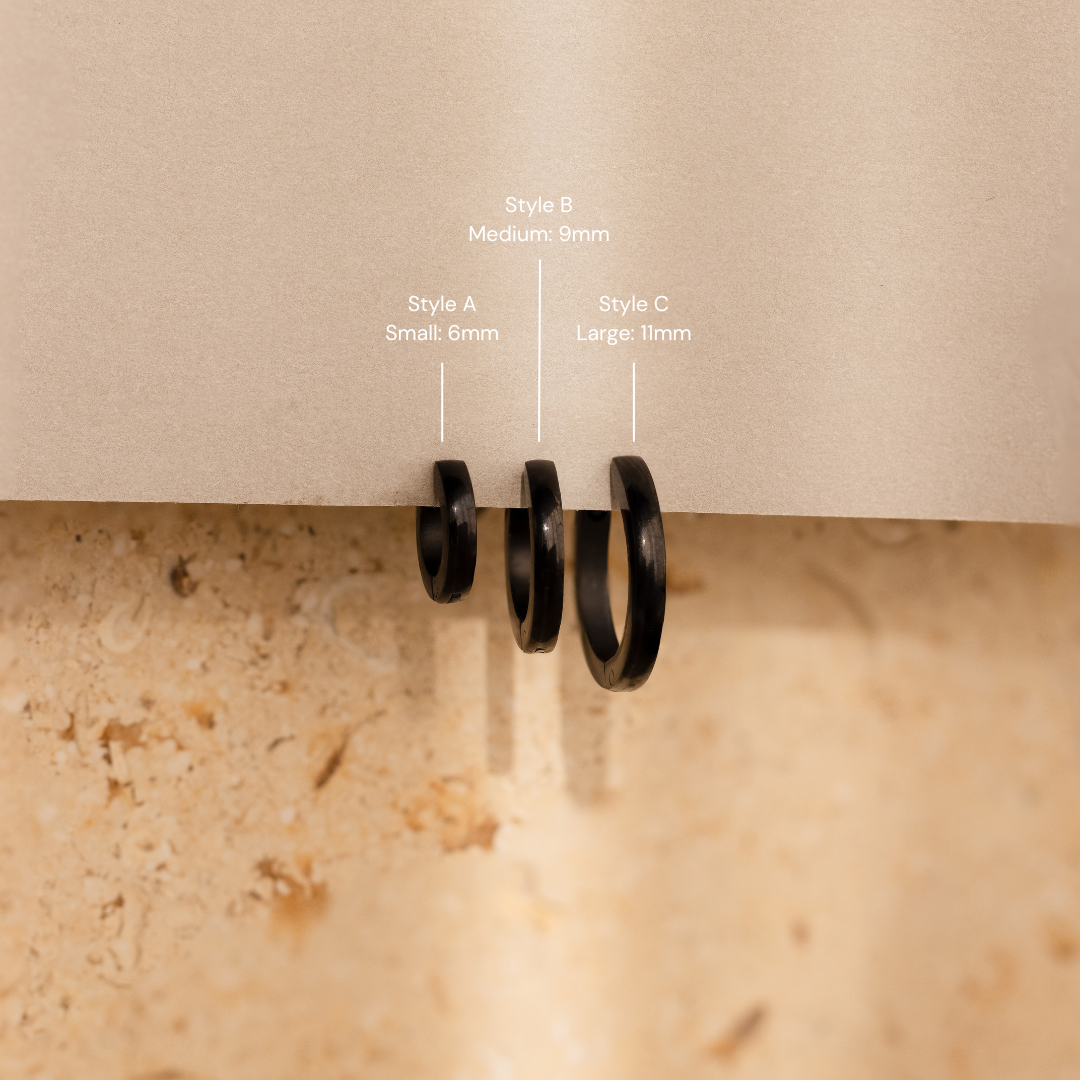 The Men's Black Hoops Set features three sleek black earrings in small, medium, and large sizes, displayed on a beige surface with labels—an ideal stylish gift for him.