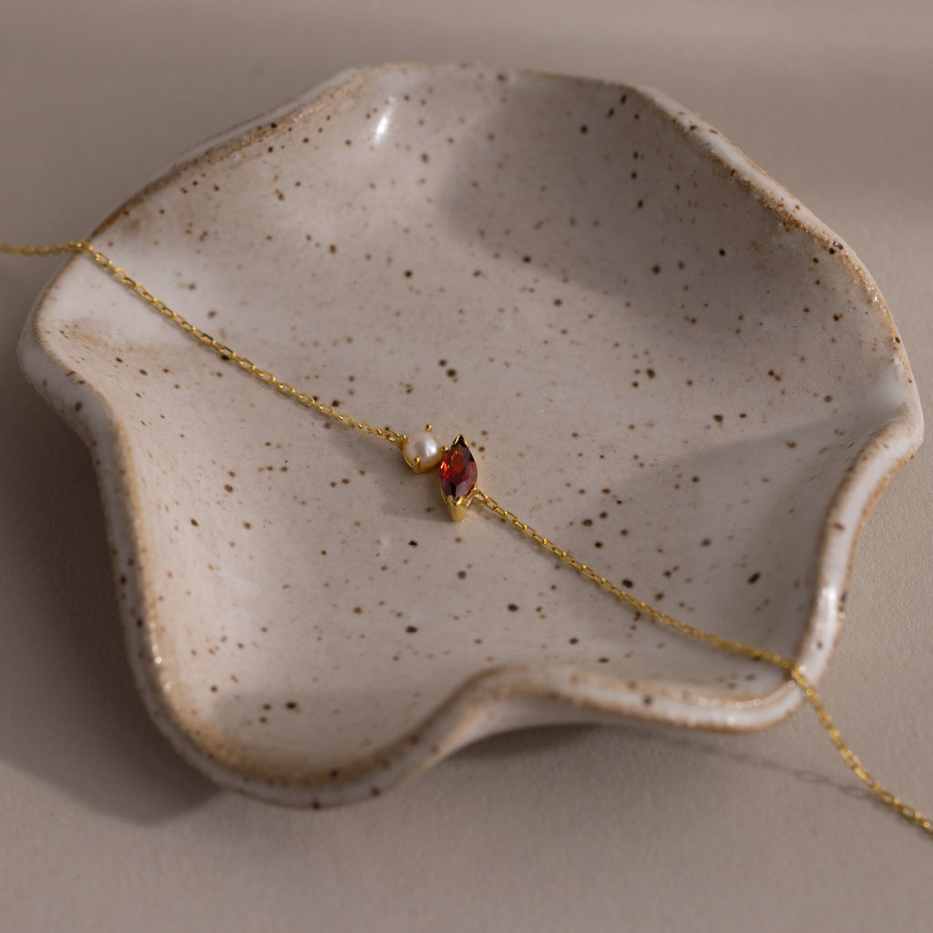 The Marquise Garnet & Pearl Bracelet, featuring a gold chain and red marquise gem, rests on a wavy-edged speckled ceramic dish—a thoughtful gift for someone special.