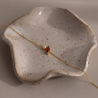The Marquise Garnet & Pearl Bracelet, featuring a gold chain and red marquise gem, rests on a wavy-edged speckled ceramic dish—a thoughtful gift for someone special.