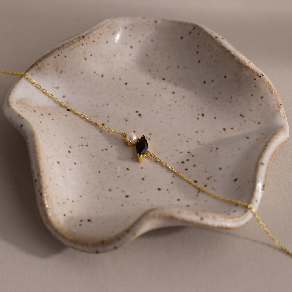 The Marquise Onyx & Pearl Bracelet, featuring a gold chain, pearls, and black onyx charm, is displayed on a speckled ceramic dish—a stunning gift to add elegance to any jewelry collection.