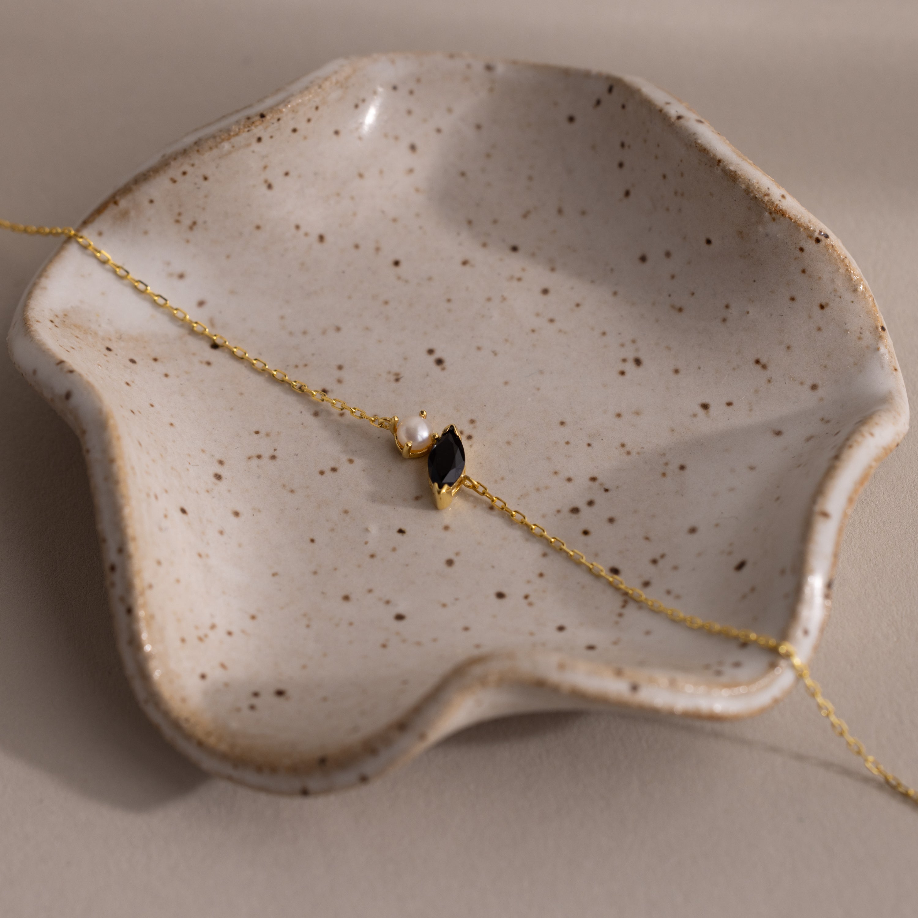 The Marquise Onyx & Pearl Bracelet, featuring a gold chain, pearls, and black onyx charm, is displayed on a speckled ceramic dish—a stunning gift to add elegance to any jewelry collection.