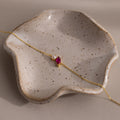 The Marquise Ruby & Pearl Bracelet, featuring a gold chain and pink stone, rests on a speckled ceramic dish—an elegant gift for someone special.