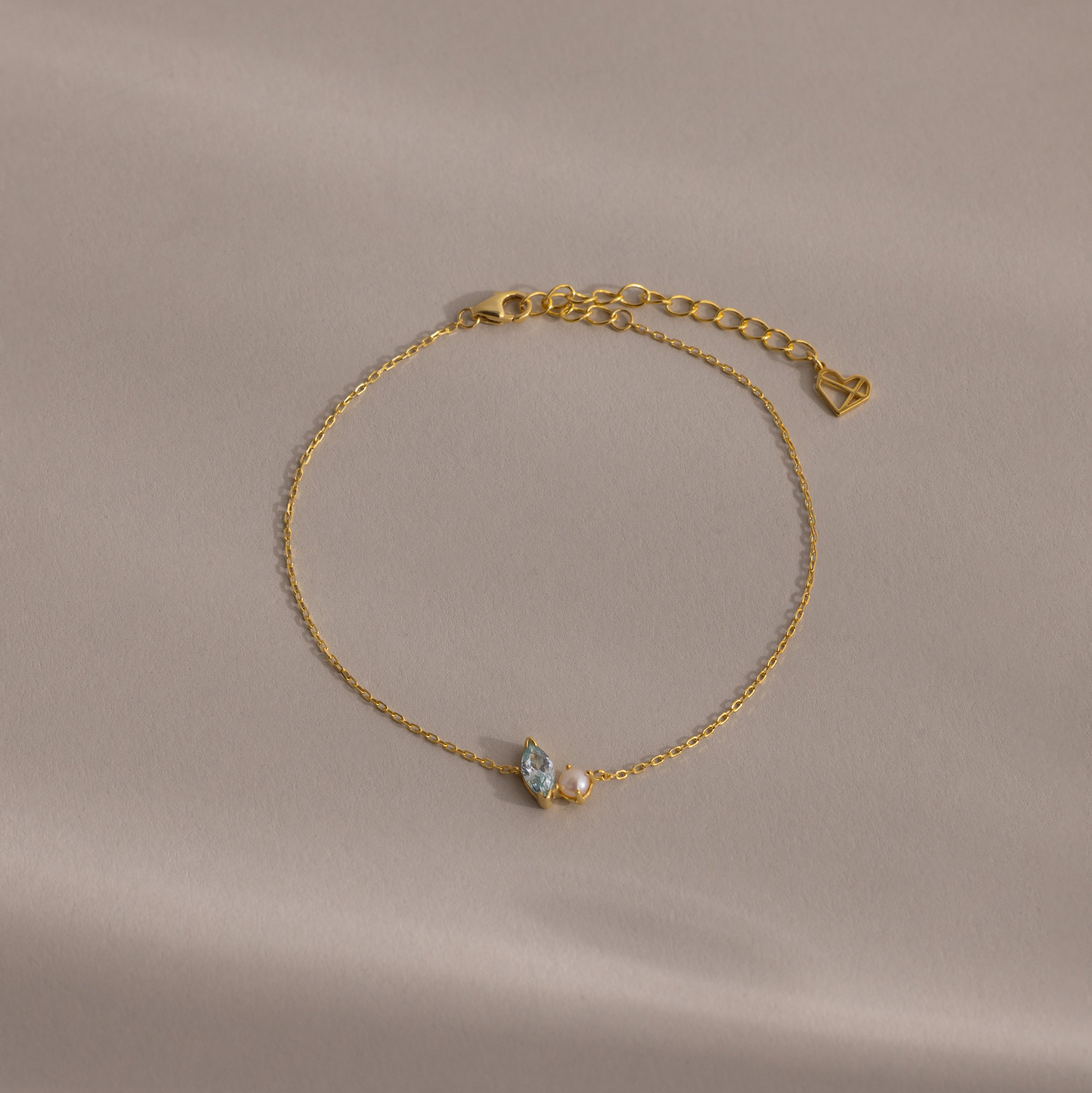 The Marquise Opal & Pearl Bracelet features two small gemstones and a heart charm, shown on a light beige background. A delicate gold bracelet—perfect as a gift for someone special.