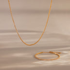 The Dainty Diamond Tennis Necklace & Bracelet Set in 18K Gold sparkles with small diamonds on a neutral beige background—an elegant choice for anniversary jewelry.
