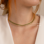 Close-up of a woman layering jewelry, wearing the Tennis Necklace Set featuring one necklace with green stones and one with white stones.