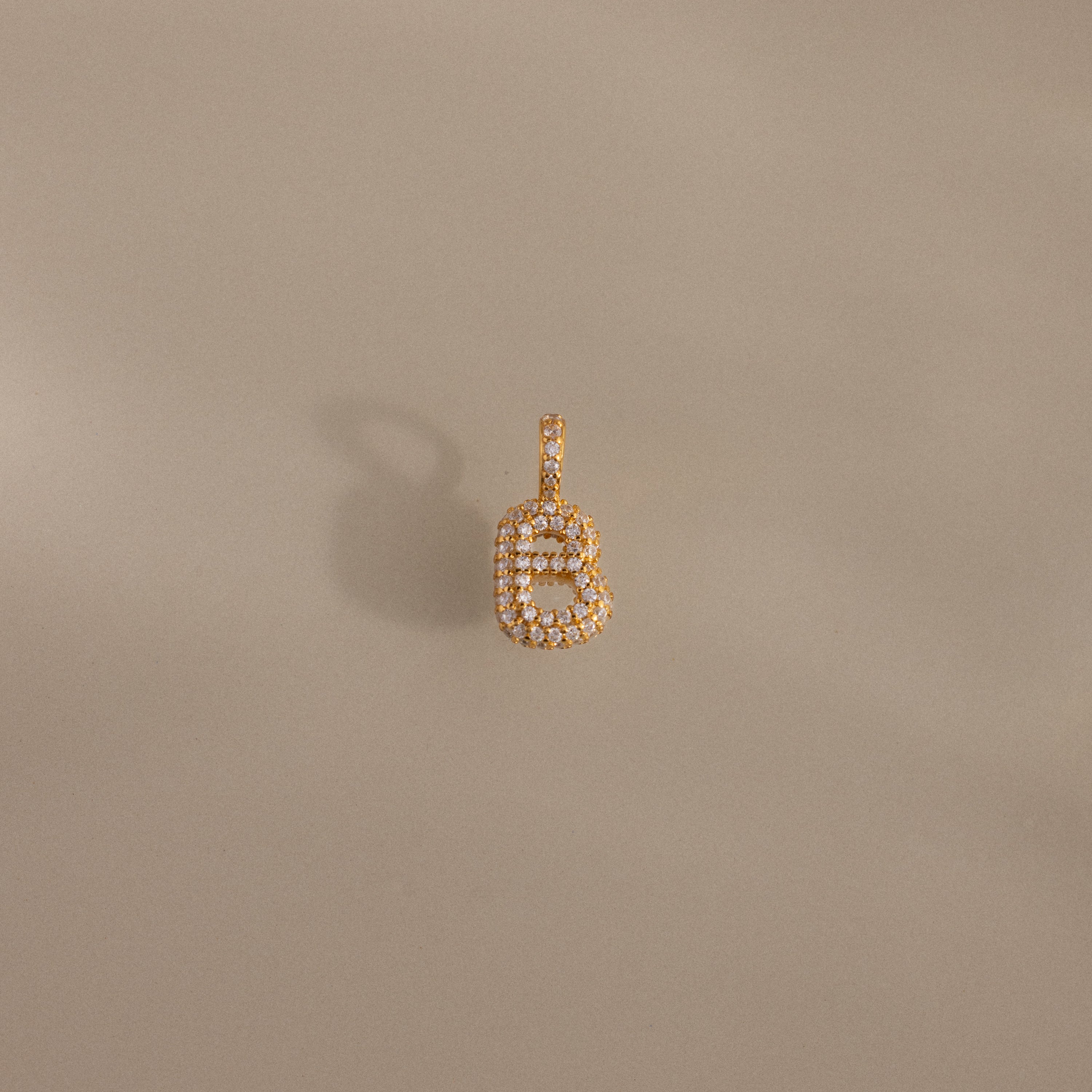 The Pave Bubble Letter Charm is a gold and diamond pendant shaped like the letter B, shown on a beige background with a soft shadow.