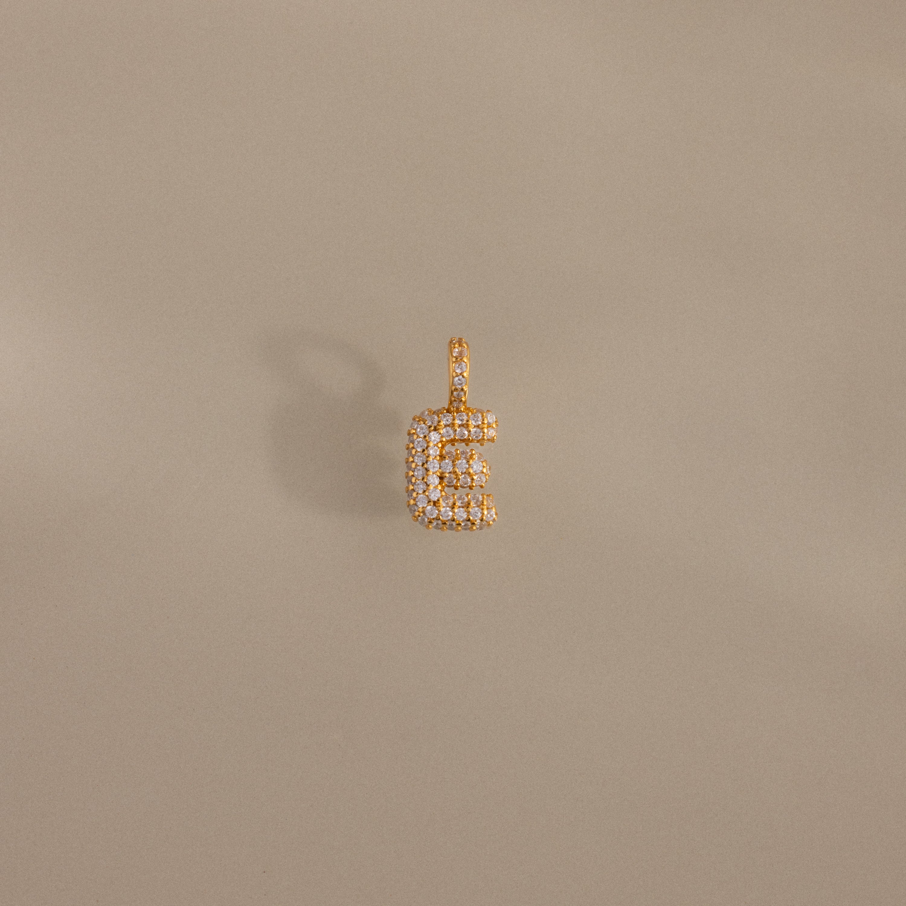 The Pave Bubble Letter Charm is a gold and diamond pendant shaped like the letter E, with diamond-studded lettering on a plain beige background.