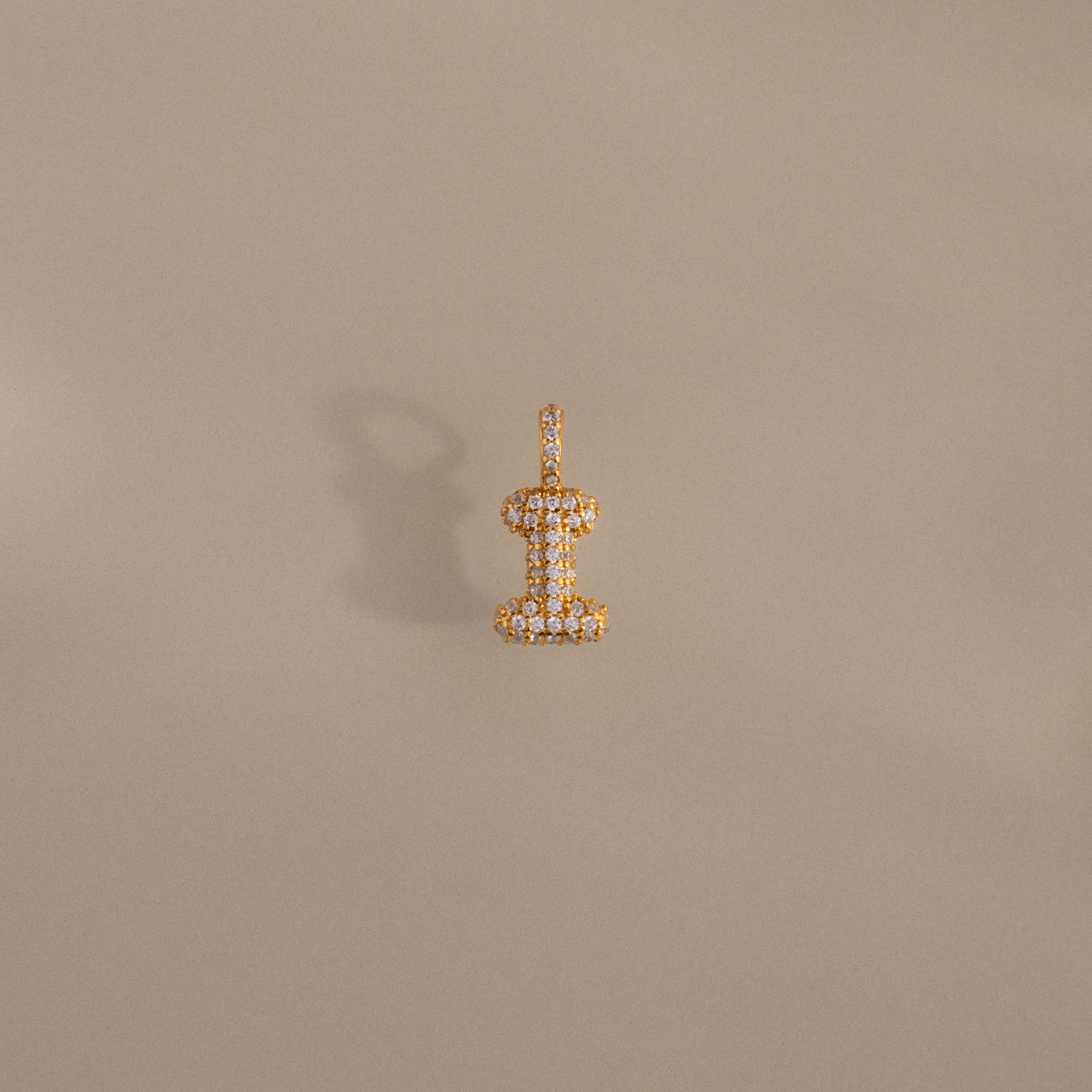 The Pave Bubble Letter Charm, shaped like a dumbbell and set against a beige background with a soft shadow, features sparkling diamond-studded lettering that enhances its elegant gold and diamond design.
