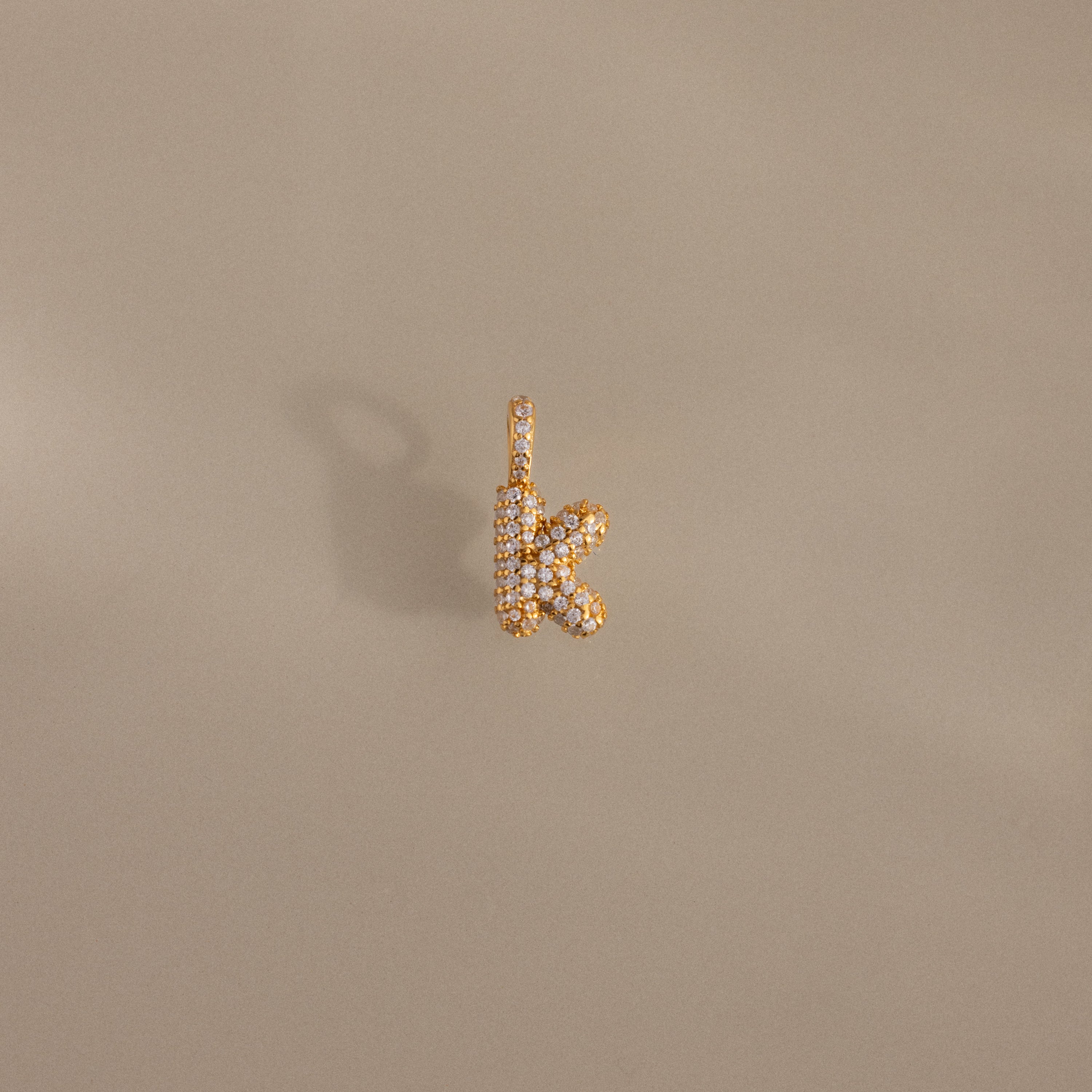 The Pave Bubble Letter Charm pendant, shaped as the letter K and adorned with diamond-studded details, is displayed against a neutral beige background.