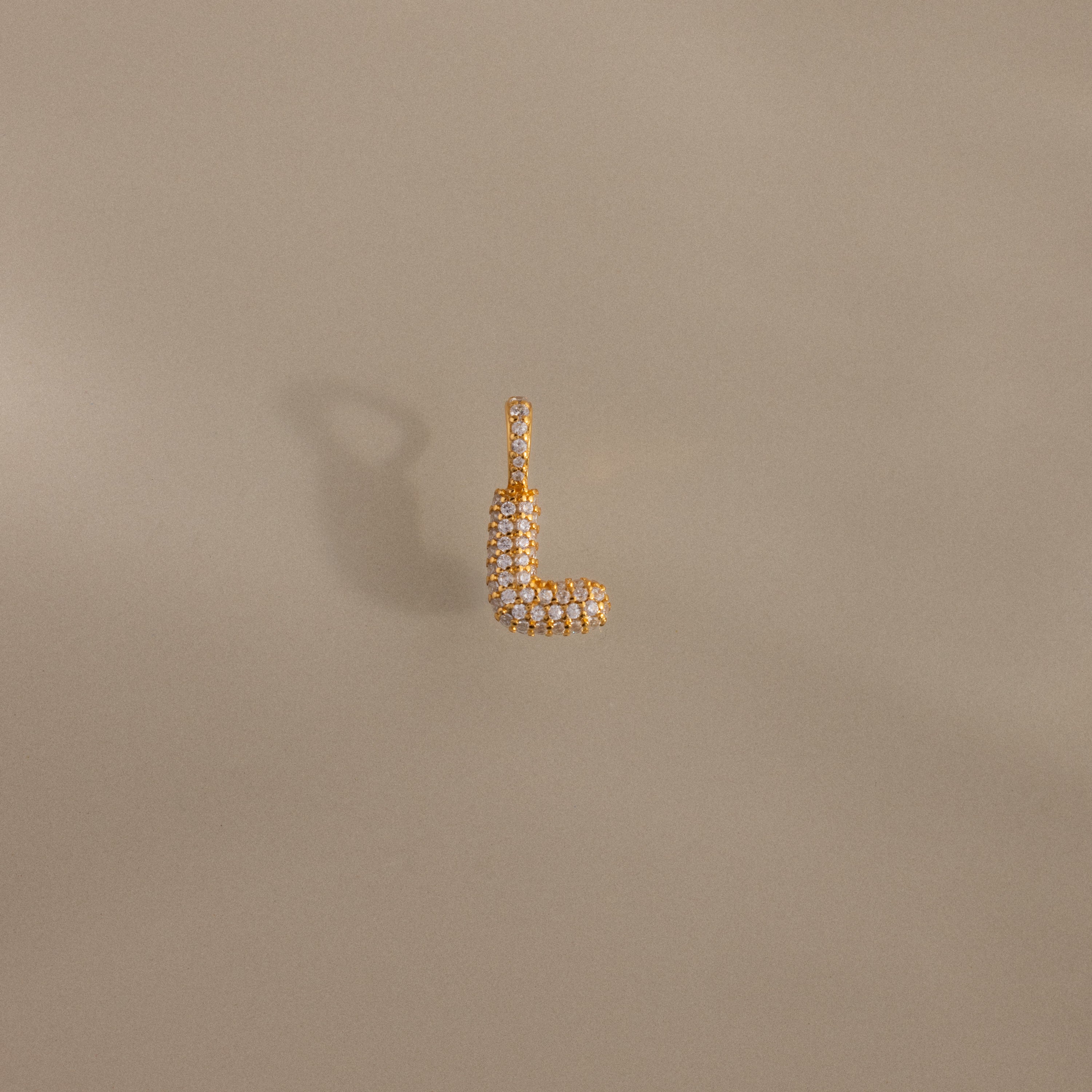 The Pave Bubble Letter Charm is a gold L-shaped charm with diamond-studded lettering in a pave bubble style, showcased on a beige background.