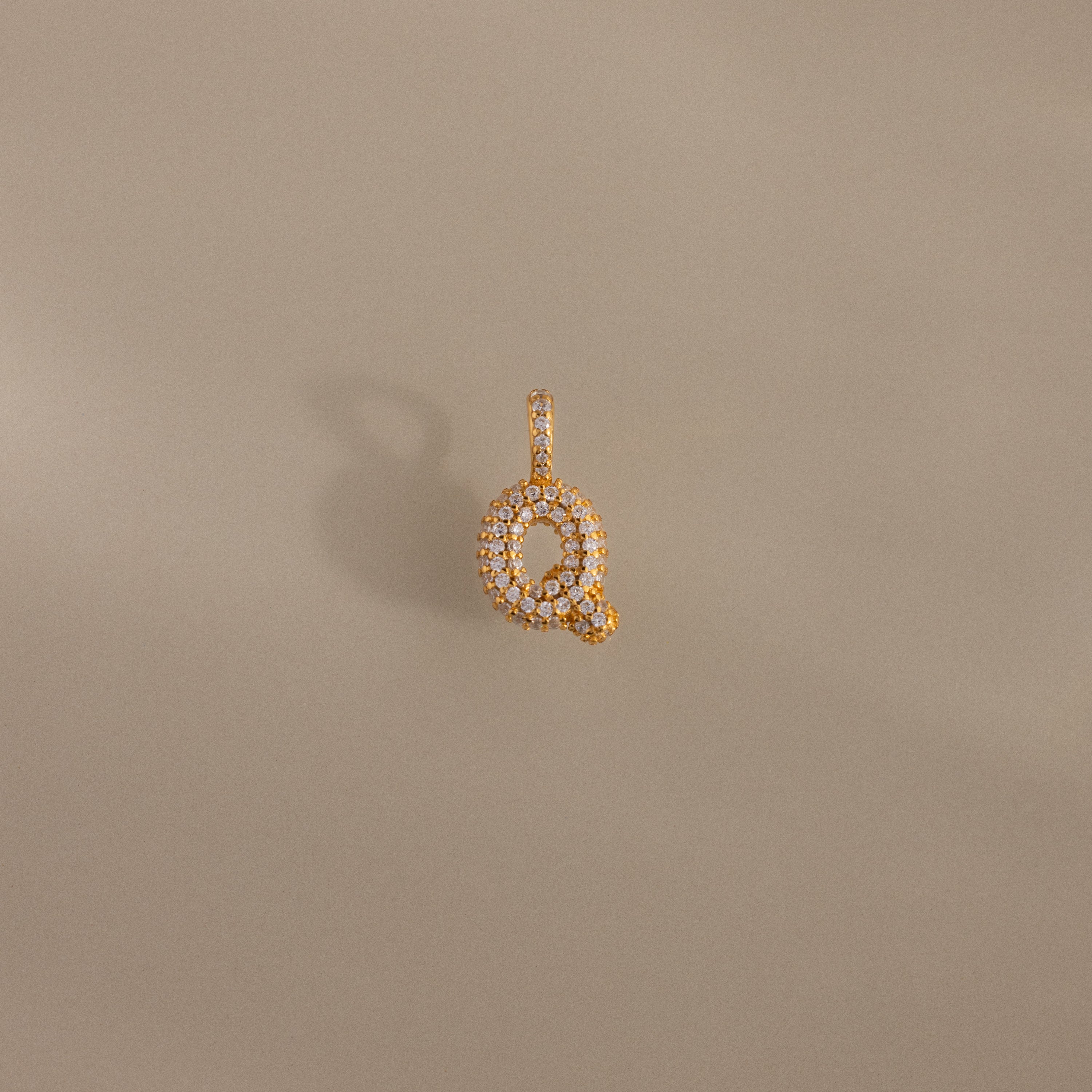 The Pave Bubble Letter Charm is a gold pendant shaped like the letter Q, featuring diamond-studded lettering with sparkling white diamonds, shown on a beige background.