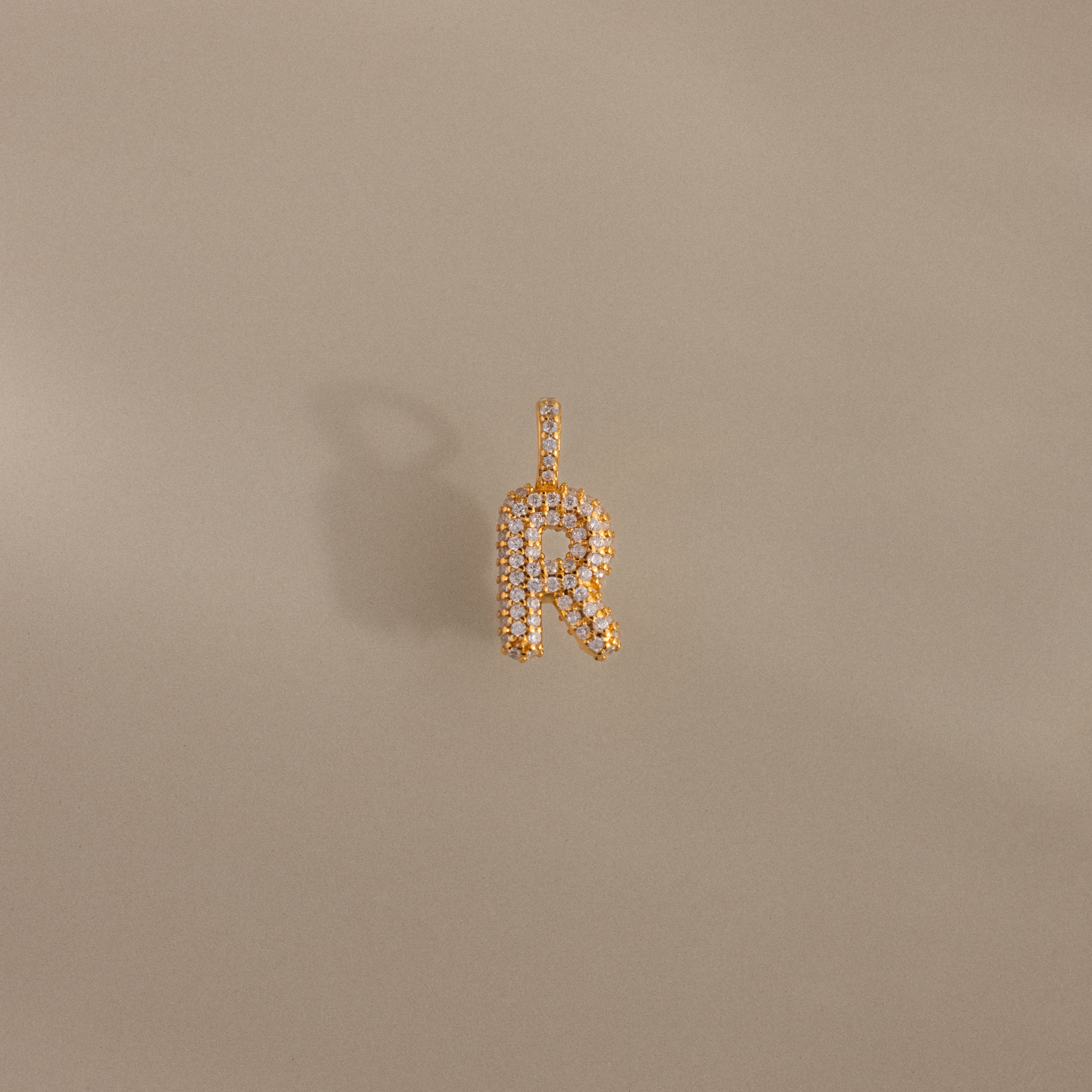 The Pave Bubble Letter Charm is a gold pendant shaped like the letter R with diamond-studded lettering and small gems, shown on a beige background.