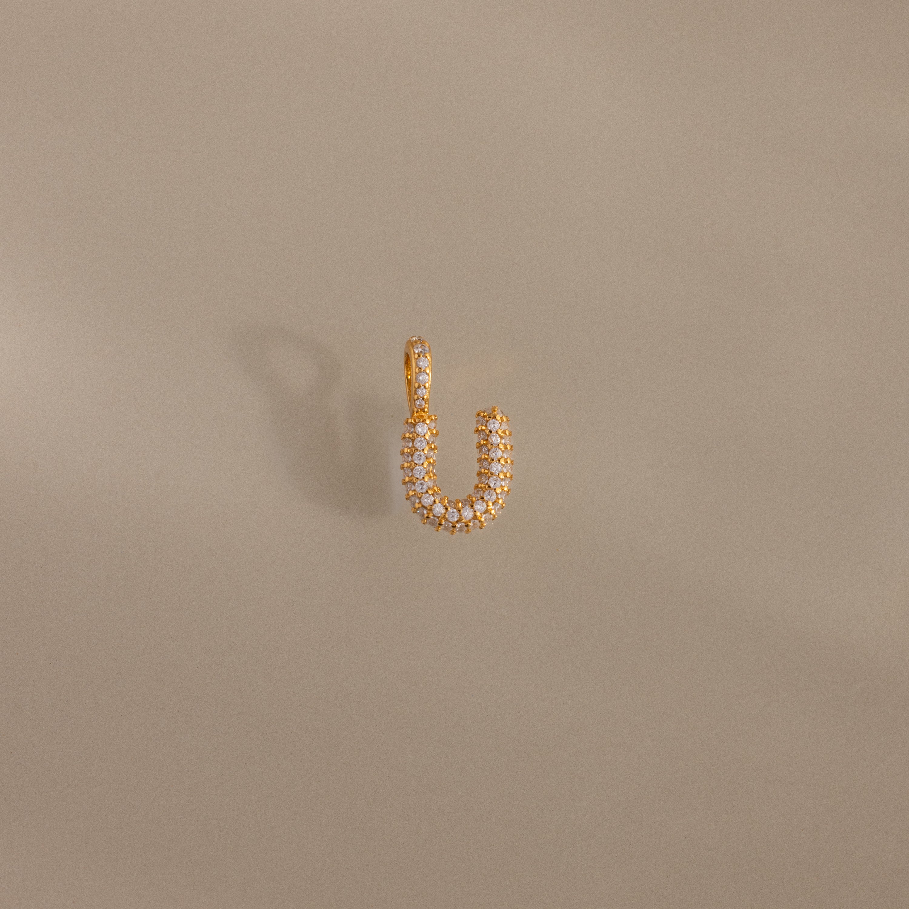 The Pave Bubble Letter Charm features a gold U-shape encrusted with small white stones, set against a neutral beige background.