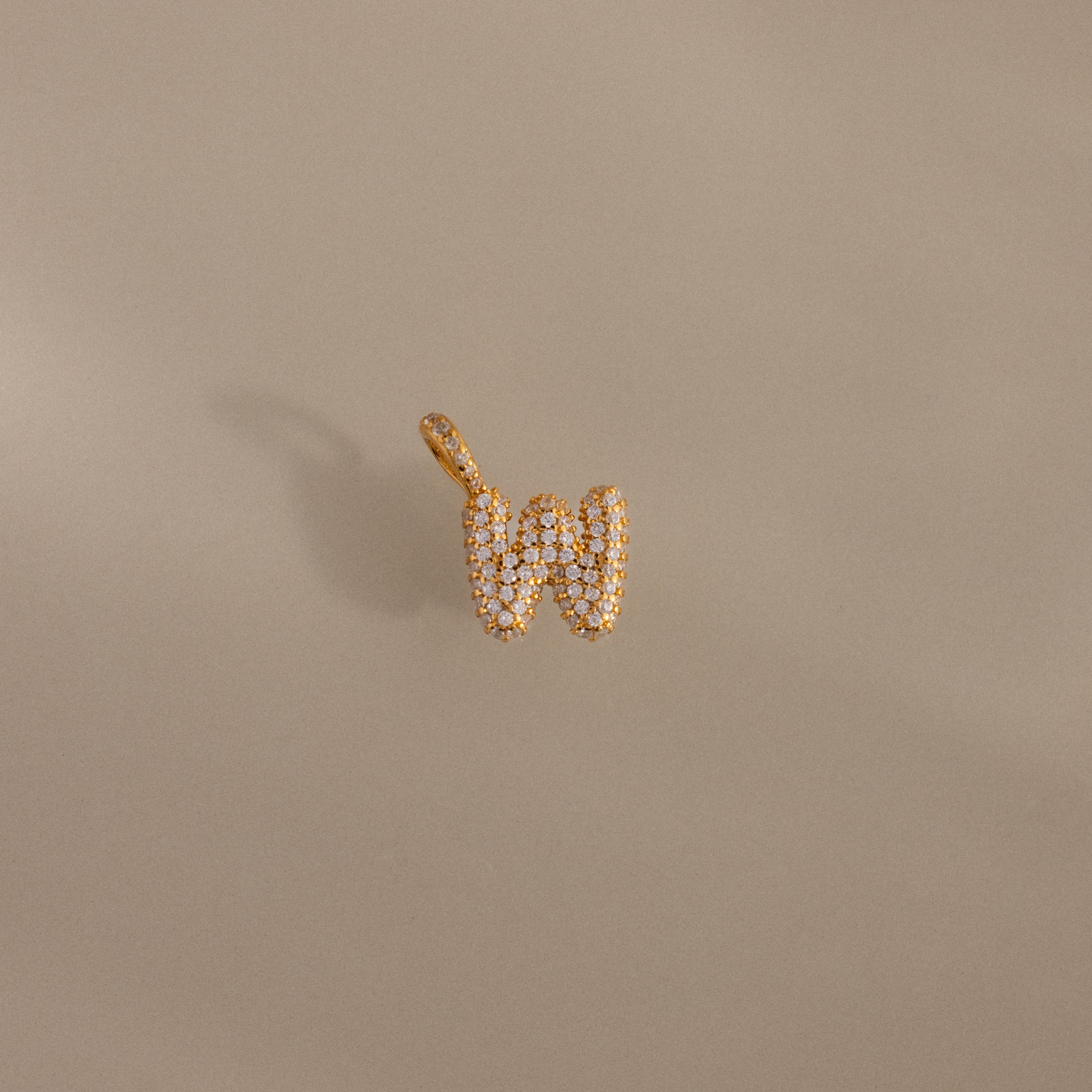 The Pave Bubble Letter Charm features gold and diamond accents in the shape of the letter W, displayed on a plain beige background.