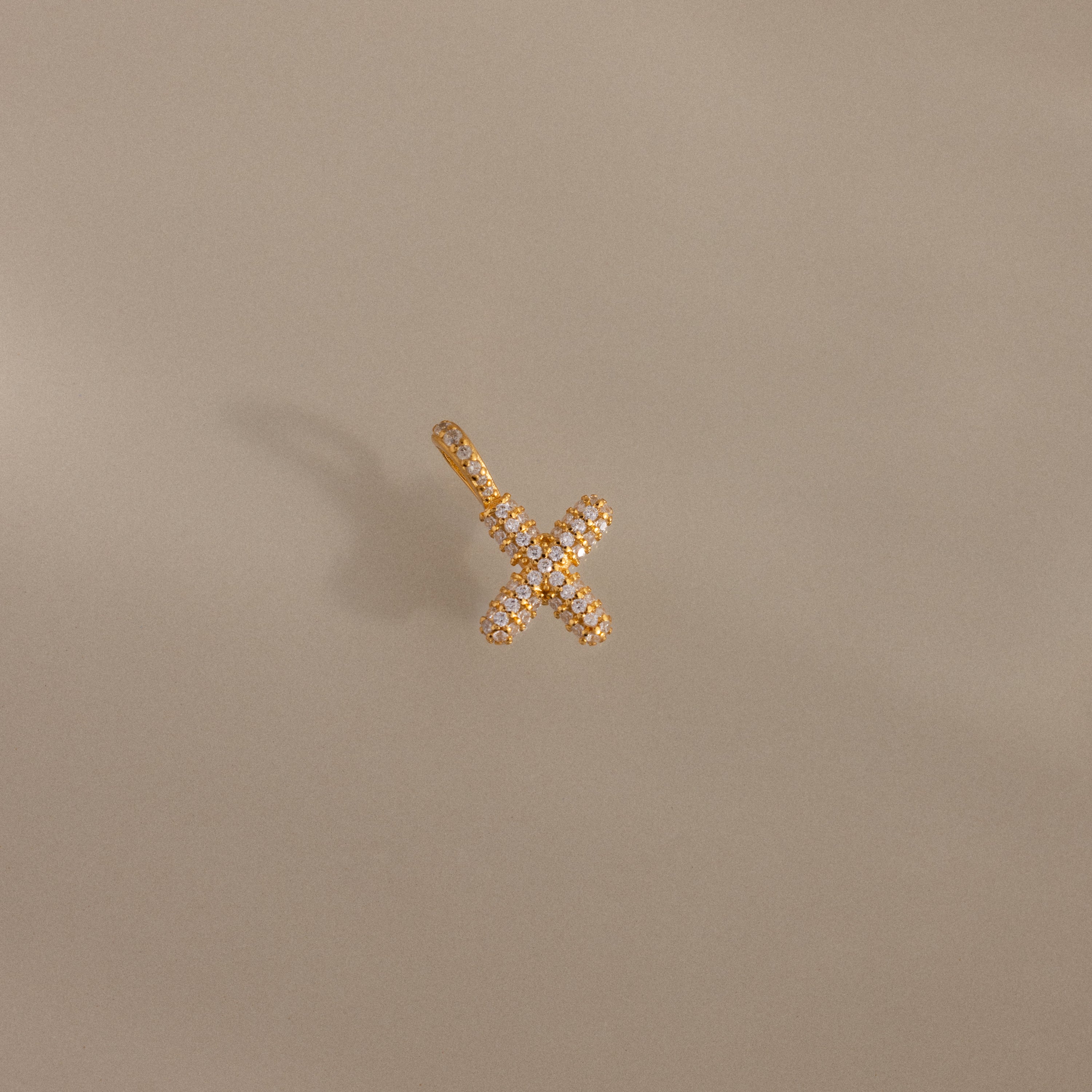 The Pave Bubble Letter Charm features a crisscross design with small diamonds and subtle diamond-studded lettering, displayed on a neutral beige background.