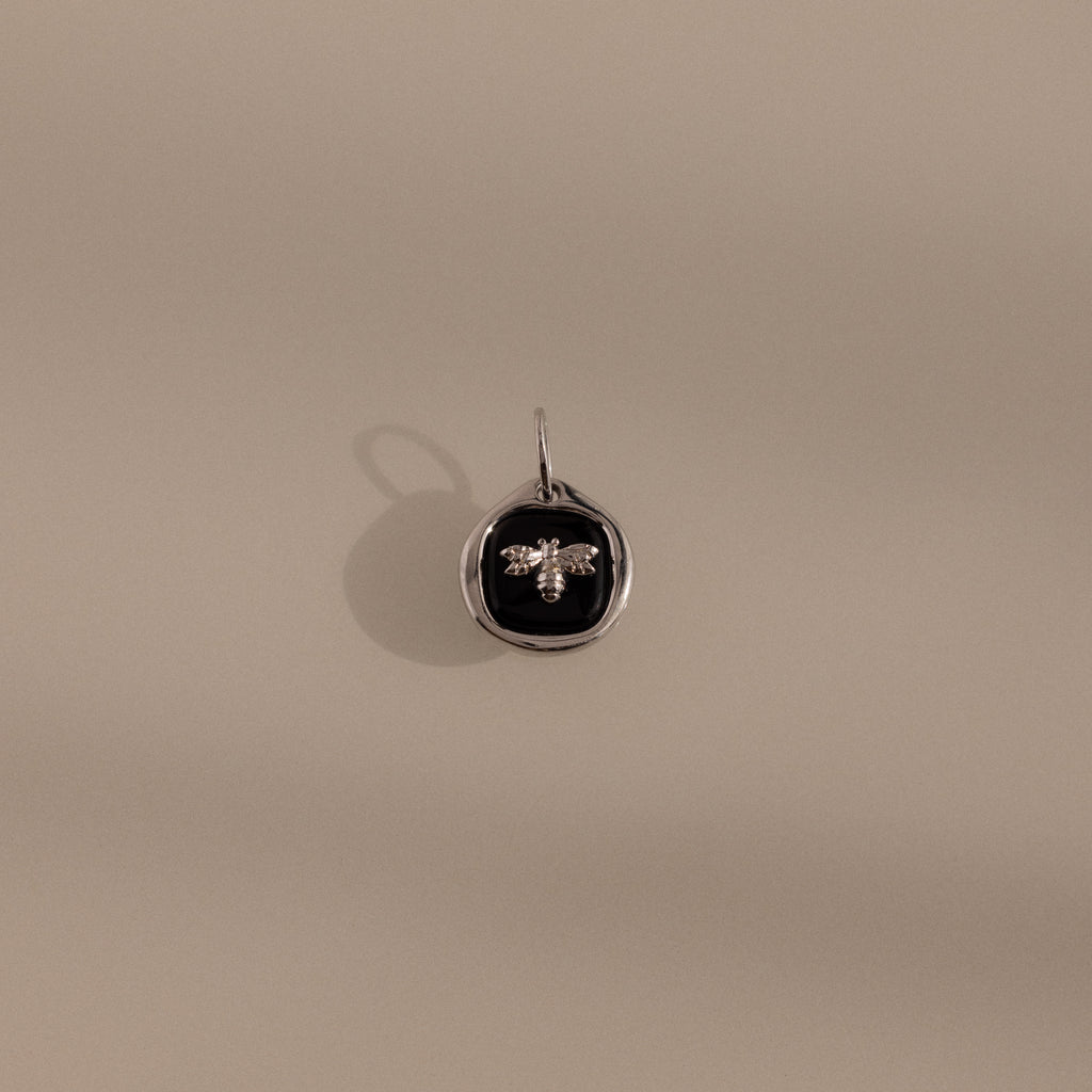 The Onyx Honeybee Charm in Silver features a raised bee on a black onyx oval, shown against a beige background—a striking accessory that effortlessly elevates your style.