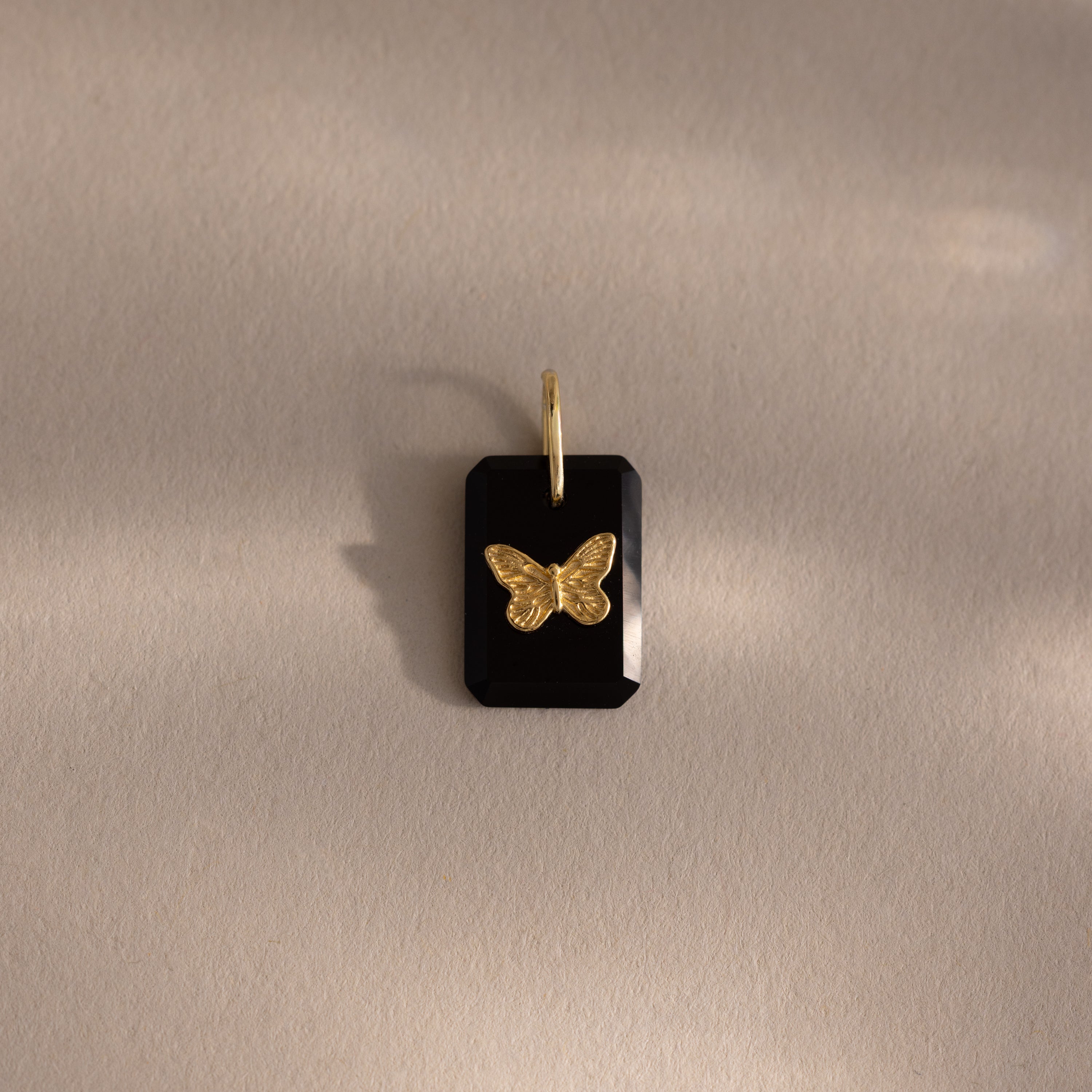 The Onyx Butterfly Charm in 18K Gold is a black rectangular pendant with a gold butterfly on a soft beige background, perfect for adding an elegant touch to any outfit.