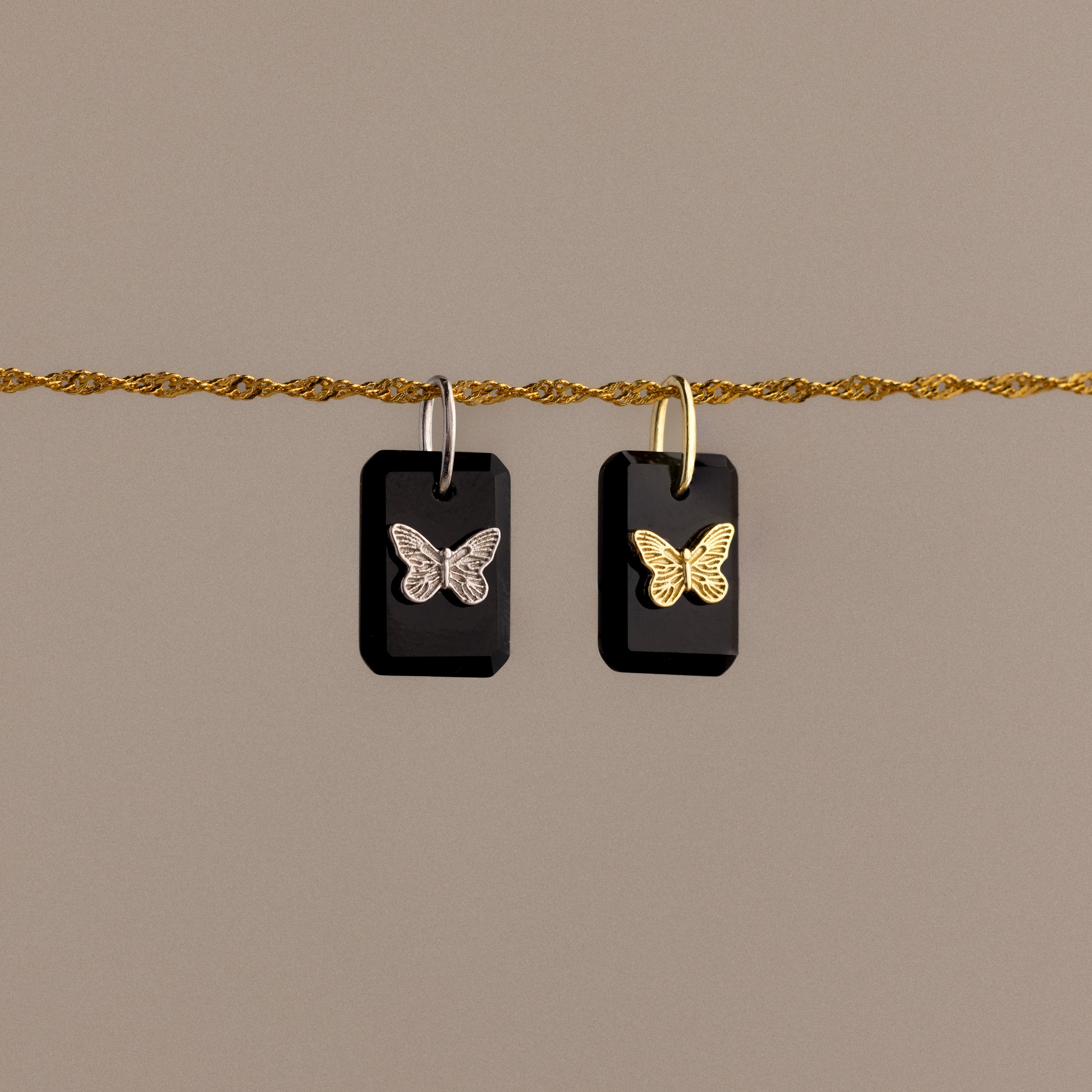 The Onyx Butterfly Charm in Silver features two black rectangular pendants with butterfly designs on a silver chain, making an elegant statement against a beige background.