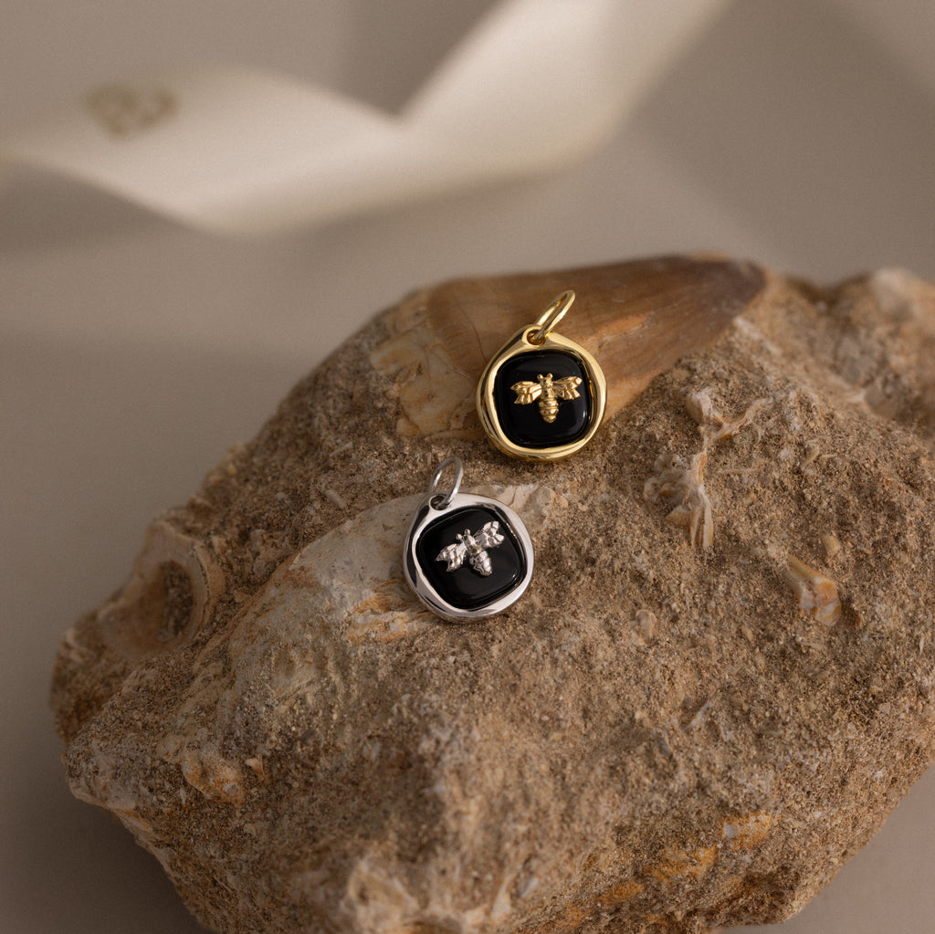 The Onyx Honeybee Charm, shown in both gold and silver on a textured rock with a neutral backdrop, brings nature-inspired charm to any jewelry collection.