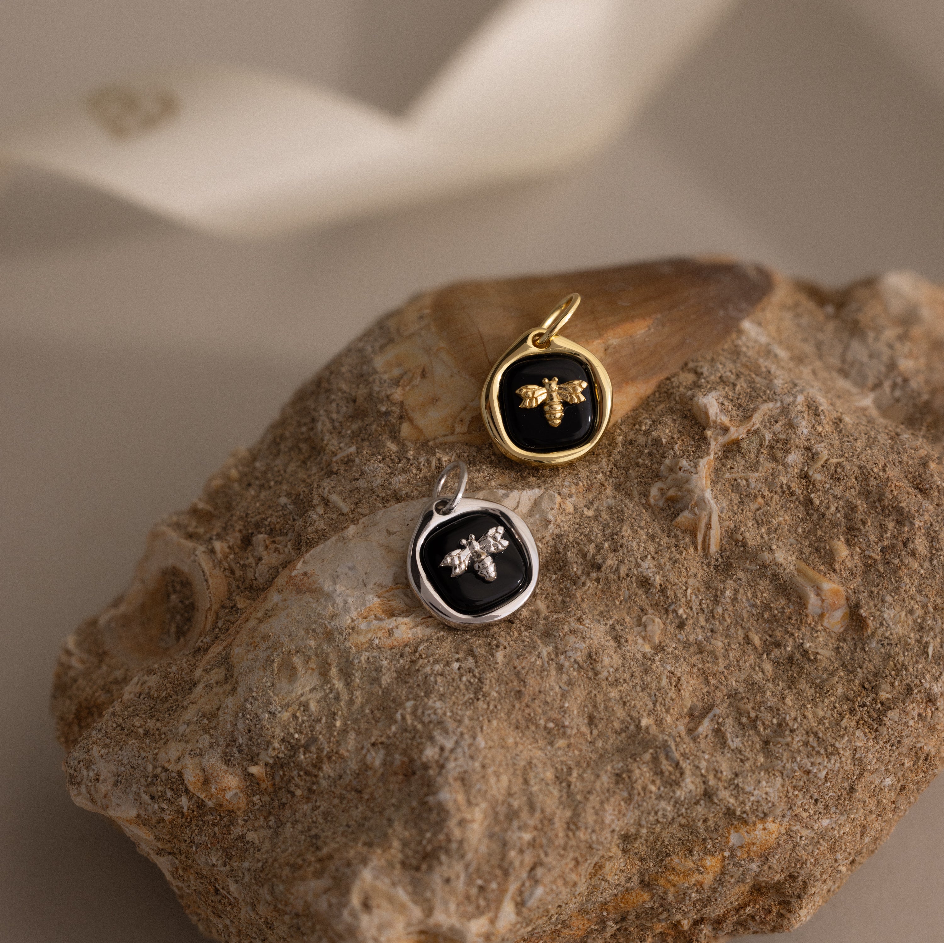 The Onyx Honeybee Charm, shown in both gold and silver on a textured rock with a neutral backdrop, brings nature-inspired charm to any jewelry collection.