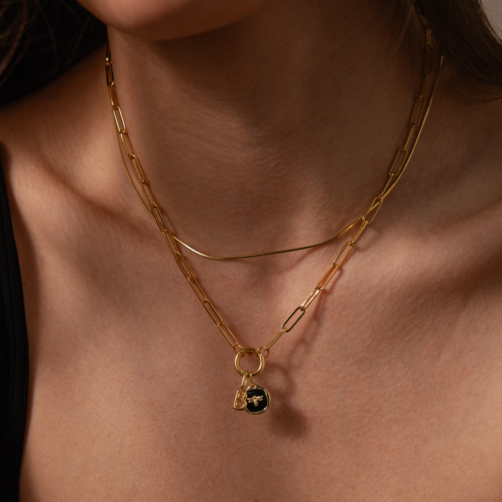 A woman wears layered gold necklaces, including the Onyx Honeybee Charm in Silver featuring a black onyx pendant and delicate charm, against bare skin.