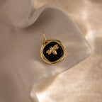 The Onyx Honeybee Charm in 18K Gold glows against a black background, displayed elegantly on a wavy cream surface.