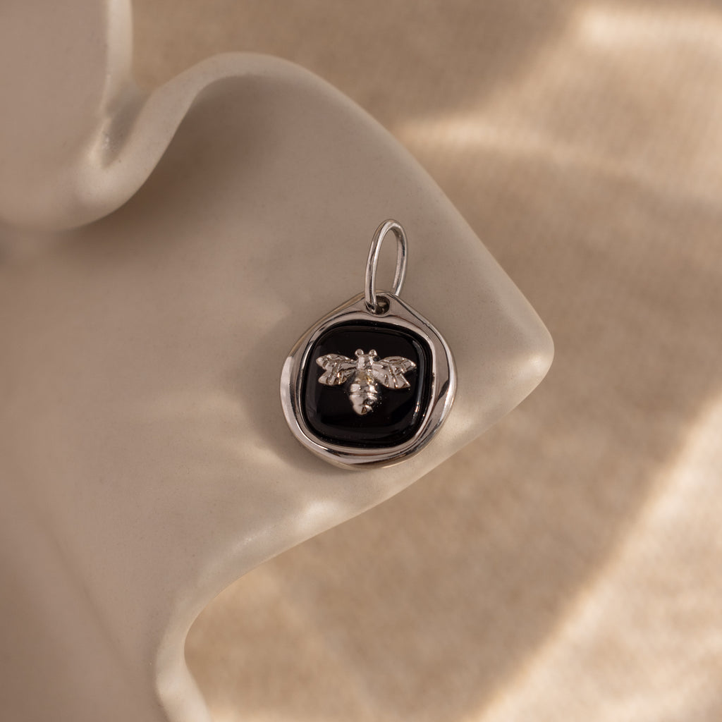 The Onyx Honeybee Charm in Silver is beautifully presented on a wavy, light background, making it a distinctive addition to any jewelry collection.